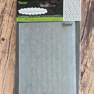 May include: A Darice Embossing Essentials embossing folder measuring 4.25 inches by 5.75 inches. The clear plastic folder features a repeating pattern of vertical arrows and small hearts, designed for creating textured impressions on paper crafts.
