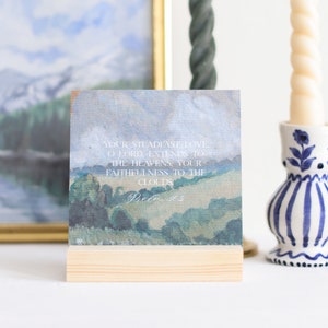 May include: A square art print with a landscape painting, mounted on a small wooden stand. The artwork features rolling green hills under a cloudy sky, with text that reads "YOUR STEADFAST LOVE O LORD, EXTENDS TO THE HEAVENS, YOUR FAITHFULNESS TO THE CLOUDS".
