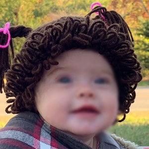 May include: A baby wearing a brown yarn wig with pink hair ties. The wig is styled in two pigtails.