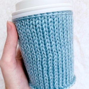 Puede incluir: Una funda de punto color azul claro envuelta alrededor de un vaso con tapa de plástico blanca. La funda presenta una textura acanalada vertical creada por puntos de tejido, proporcionando un ajuste ceñido alrededor del cuerpo cilíndrico.