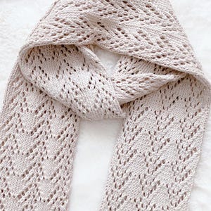May include: A light beige knitted scarf with a delicate openwork design. The scarf is folded, showcasing the intricate pattern of small holes and chevron-like shapes. The soft, neutral color makes it a versatile accessory.