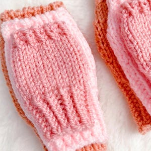 May include: A pair of fingerless gloves featuring a layered design with a light pink ribbed knit section centered over a peach-colored base. The gloves are constructed with a vertical knit pattern and finished with horizontal ribbed cuffs at the edges.