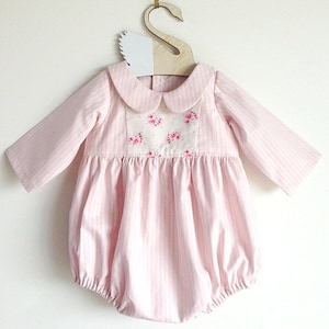 May include: A pink and white striped baby onesie with a white collar and a floral design on the front. The onesie has long sleeves and a bubble hem.