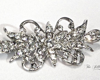 May include: A silver-toned brooch with a floral design, featuring a large central flower with smaller flowers and leaves surrounding it. The brooch is adorned with clear rhinestones.