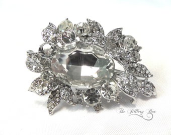 May include: Silver-toned brooch with a large, clear, oval-cut rhinestone as the focal point. The brooch features a floral design with smaller, round rhinestones and intricate metalwork, creating a sparkling, elegant accessory.
