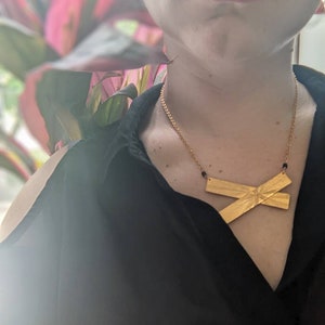 May include: A person wearing a black shirt with a gold necklace featuring a large, textured, X-shaped pendant.