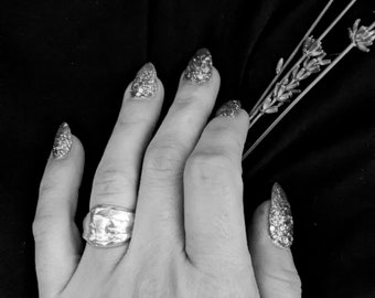 May include: A hand with silver glitter nail polish and a silver ring on the middle finger. The hand is resting on a black fabric background.