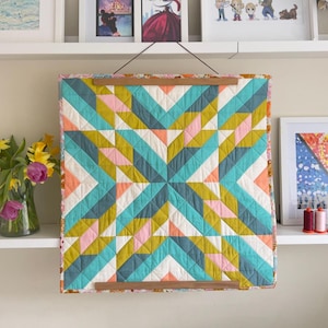 May include: A geometric quilt with a starburst pattern, using teal, pink, yellow, and white triangles. The quilt is displayed on a wooden hanging frame. The quilt is a decorative item.