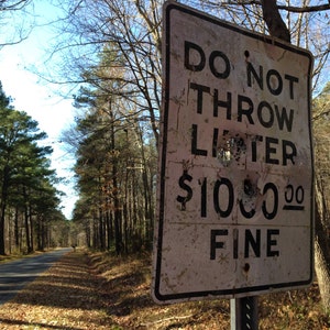 May include: A white sign with black text reads "Do Not Throw Litter $100.00 Fine". The sign is weathered and has several bullet holes. The sign is in a wooded area with trees and a paved road in the background.