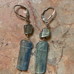 Op de afbeelding: Een paar zilveren oorhangers met blauwe en grijze edelsteenaccenten. De oorbellen hebben een lange, rechthoekige blauwe edelsteen, een kleinere grijze edelsteen en een zilveren haak.