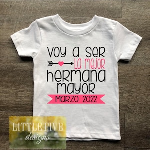 May include: White t-shirt with black text that reads "Voy a ser la mejor hermana mayor Marzo 2022". The text is decorated with a pink heart and arrow.