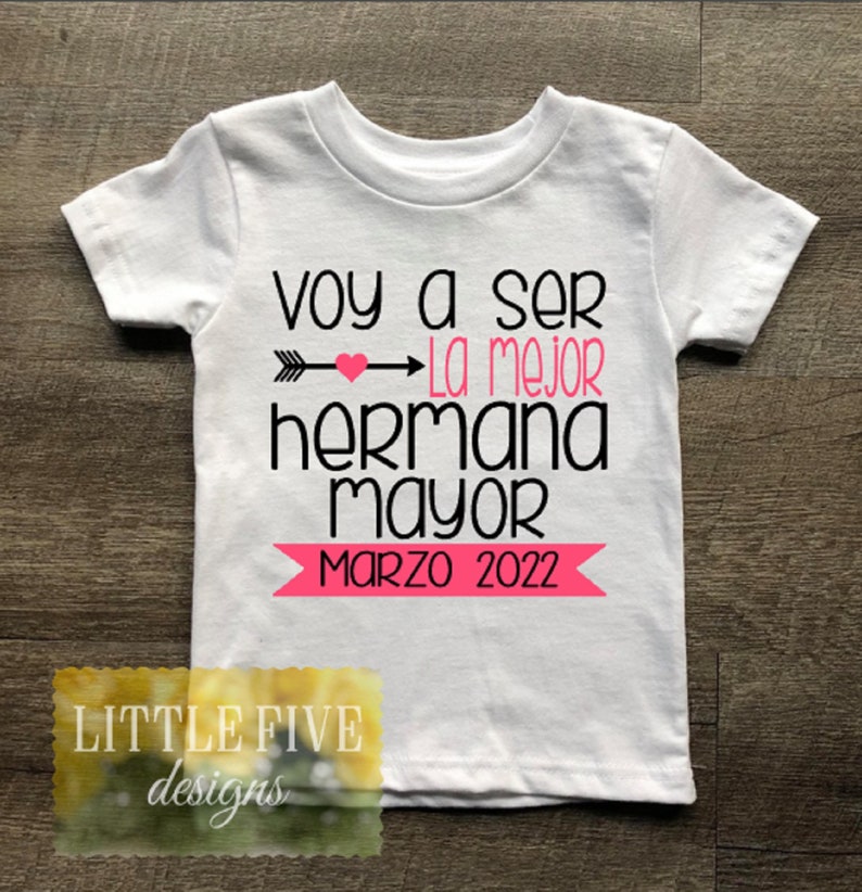 May include: White t-shirt with black text that reads "Voy a ser la mejor hermana mayor Marzo 2022". The text is decorated with a pink heart and arrow.