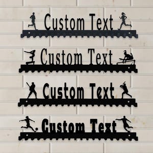 Peut inclure: Présentoirs de médailles de sport en métal noir avec les mots "Custom Text". Chaque présentoir présente des silhouettes d'athlètes dans divers sports : course, haies, baseball et football. Les présentoirs sont montés sur un mur de briques blanches.