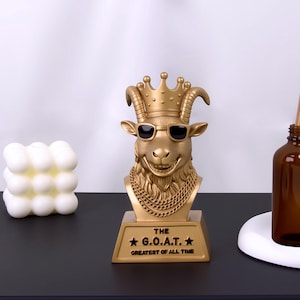 Op de afbeelding: Een goudkleurige geitenbust met zonnebril en kroon. De buste heeft een kettingketting en is gemonteerd op een voet met de tekst "THE G.O.A.T. GREATEST OF ALL TIME". Een decoratief item.