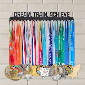 以下が含まれることがあります： 「DREAM. TRAIN. ACHIEVE」の文字が書かれた黒い金属製のメダルディスプレイ。カラフルなリボンとメダルが並んでいます。メダルには「RUN」や「Shanghai Online Race」など、さまざまなデザインとテキストが施されています。