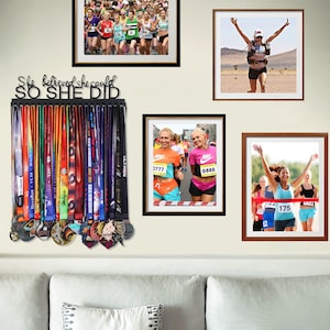 Puede incluir: Un soporte para medallas de metal negro con el texto "She believed she could SO SHE DID". El soporte est&aacute; lleno de medallas y cintas de colores. Hay fotos enmarcadas de corredores en la pared detr&aacute;s del soporte.