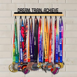Pode incluir: Um expositor de medalhas de madeira com as palavras "DREAM. TRAIN. ACHIEVE" em preto. Fitas coloridas pendem do expositor, segurando várias medalhas. As medalhas têm diferentes designs e cores, e as fitas têm nomes de eventos.