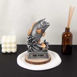 Peut inclure: Un trophée de poisson argenté et doré avec le texte "BIG FISH" sur une base en bois. Le poisson saute hors de l'eau. Une assiette blanche et ronde, une bouteille en verre marron avec des roseaux et une bougie cube blanche sont également dans l'image.