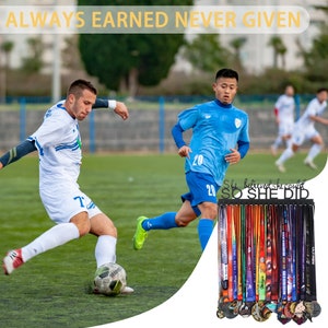 Puede incluir: Un soporte de medallas de metal negro con el texto "She believed she could SO SHE DID". El soporte est&aacute; lleno de medallas y cintas de colores. El fondo muestra un campo de f&uacute;tbol con dos jugadores con uniformes azul y blanco.