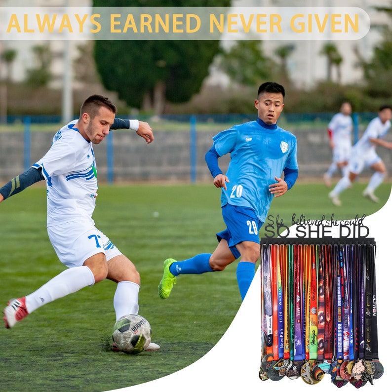 Puede incluir: Un soporte de medallas de metal negro con el texto "She believed she could SO SHE DID". El soporte est&aacute; lleno de medallas y cintas de colores. El fondo muestra un campo de f&uacute;tbol con dos jugadores con uniformes azul y blanco.