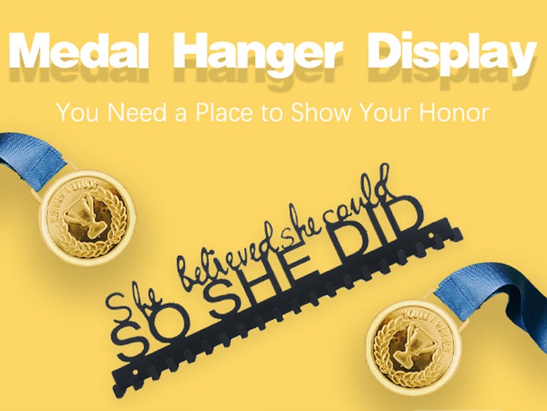 Puede incluir: Colgador de medallas de metal negro con el texto "She believed she could so she did". El colgador tiene 10 ganchos para medallas. Se muestran dos medallas doradas con cintas azules colgadas de los ganchos.