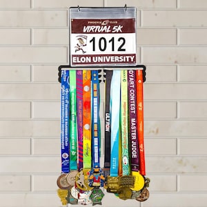 Könnte beinhalten: Ein schwarzer Medaillen-Display-Rack mit bunten Rennmedaillen und Bändern. Oberhalb des Racks befindet sich ein Schild mit der Aufschrift "PHOENIX CLUB VIRTUAL 5K 1012 ELON UNIVERSITY". Die Medaillen sind in verschiedenen Farben und Designs gehalten.