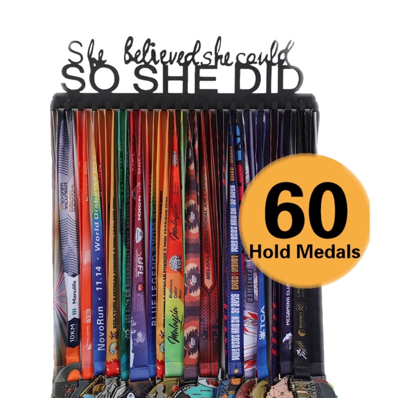 Puede incluir: Portamedallas de metal negro con el texto "She believed she could SO SHE DID". El portamedallas tiene 60 ganchos para colgar medallas. Un c&iacute;rculo amarillo con el texto "60 Hold Medals" est&aacute; en la esquina inferior derecha de la imagen.