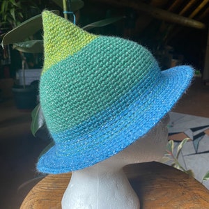 May include: A green, blue, and yellow crocheted witch hat with a pointed crown. The hat is on a white styrofoam head.