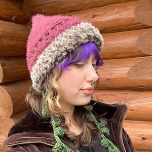 May include: A burgundy and brown knit hat with a faux fur brim. The hat is worn by a person with purple hair and a nose ring.