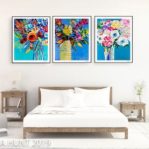 May include: Three framed abstract paintings of colorful flower bouquets in vases against a blue background. The paintings are hung on a white wall above a white bed with white bedding and a wooden frame.