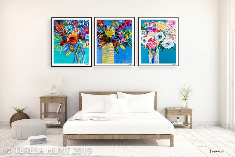 May include: Three framed abstract paintings of colorful flower bouquets in vases against a blue background. The paintings are hung on a white wall above a white bed with white bedding and a wooden frame.