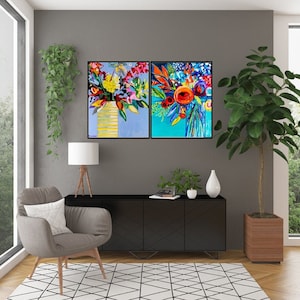 May include: Two framed abstract paintings of colorful flower arrangements. The paintings are hung on a gray wall above a black cabinet with a wooden top. A gray armchair with a white pillow sits in the corner of the room.