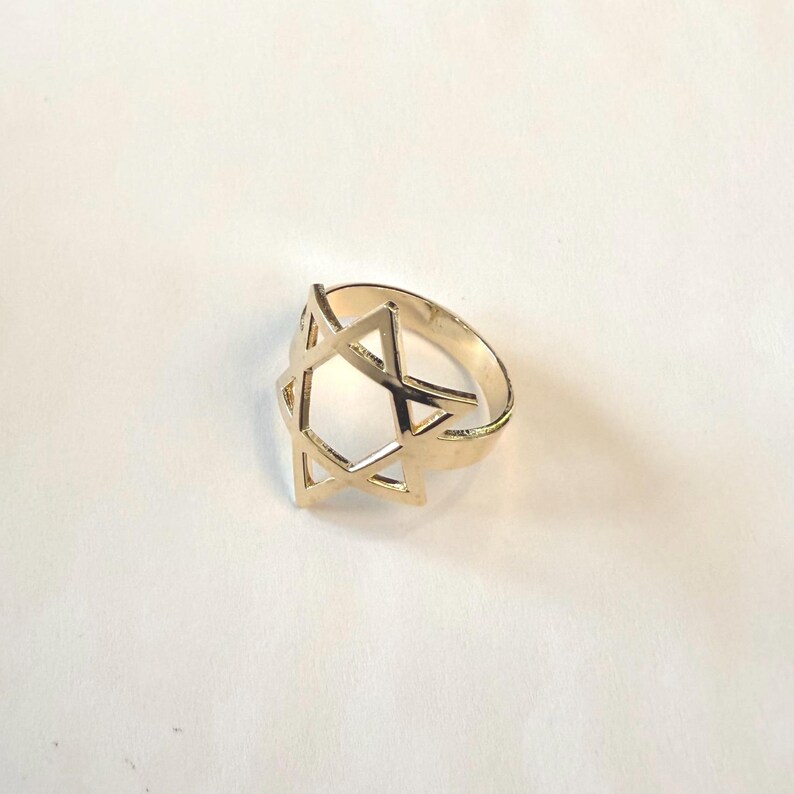 May include: A gold-colored ring featuring a Star of David design. The ring has a band and a six-pointed star, with the star's points connected to the band. The ring is set against a white background.