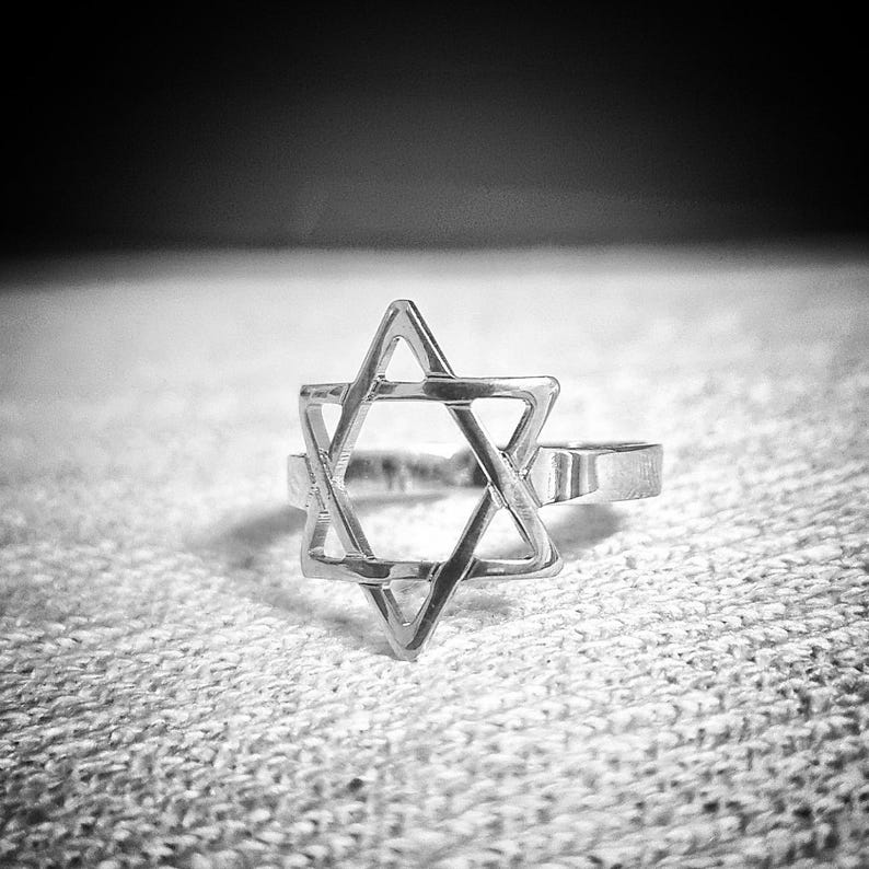 May include: A silver-colored ring featuring a Star of David design. The ring is set against a textured, light-colored surface. The six-pointed star is the focal point, with a simple band.