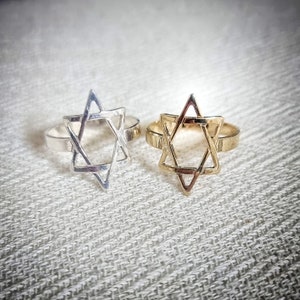 May include: Two rings, one silver and one gold, each featuring a Star of David design. The rings are set against a textured, off-white background. The Star of David is a six-pointed star, a symbol of Judaism.