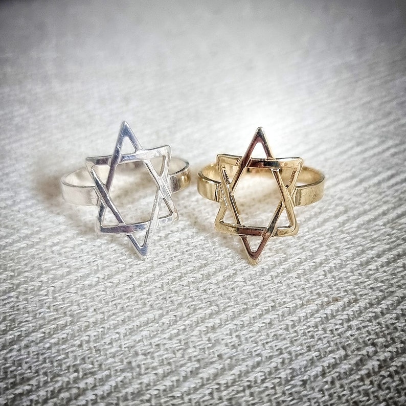 May include: Two rings, one silver and one gold, each featuring a Star of David design. The rings are set against a textured, off-white background. The Star of David is a six-pointed star, a symbol of Judaism.