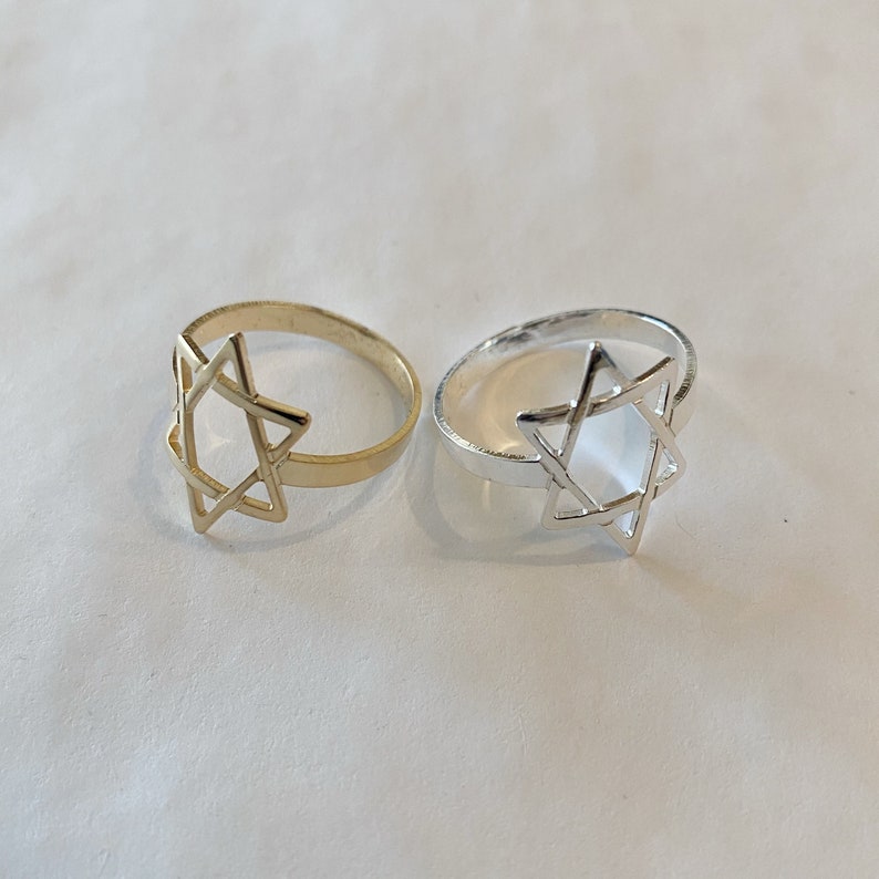May include: Two rings, one gold-toned and one silver-toned, each featuring a Star of David design. The rings are set against a plain, light-colored background. The Star of David is a six-pointed star, a prominent symbol in Judaism.
