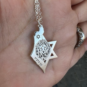 Op de afbeelding: Zilverkleurige hangerketting met een kaartomtrek, een Davidster en een leeuwenkop. De hanger bevat Hebreeuwse tekst en hangt aan een fijne ketting. De ketting wordt in een hand gehouden.