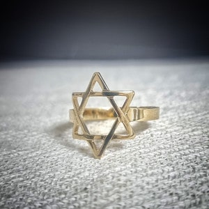 May include: A gold-colored ring featuring a Star of David design. The ring's openwork design showcases the six-pointed star, a symbol of Judaism. The ring is set against a textured, light-colored background.
