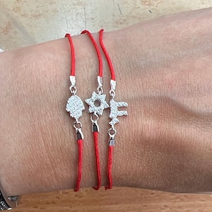 May include: Three red string bracelets with silver charms. The charms include a Hamsa hand, a star, and the letter F. The bracelets are displayed on a wrist.