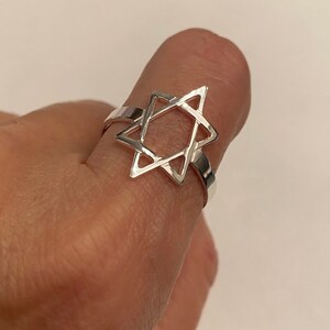 May include: A silver ring featuring a Star of David design. The ring is made of a shiny, reflective metal and is worn on a finger. The Star of David is the central focus of the ring's design.