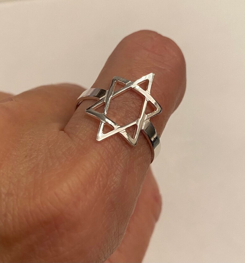 May include: A silver ring featuring a Star of David design. The ring is made of a shiny, reflective metal and is worn on a finger. The Star of David is the central focus of the ring's design.