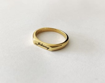 May include: A gold-colored signet ring with the name "Johnny" engraved in black script. The ring has a rectangular face and a polished finish, set against a white background. The ring is a classic piece of jewelry.