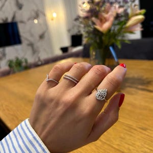 Può includere: Una mano mostra quattro anelli, ognuno con un design diverso. Gli anelli sono color argento e adornati con piccole pietre scintillanti. Le unghie sono dipinte di rosso. Lo sfondo include un tavolo di legno e un vaso di fiori.