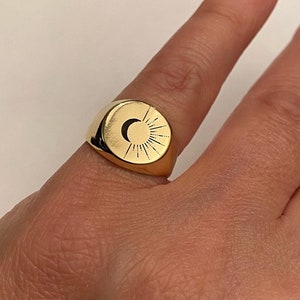 Puede incluir: Un anillo de sello de oro con un diseño de sol y luna. El anillo se lleva en un dedo.