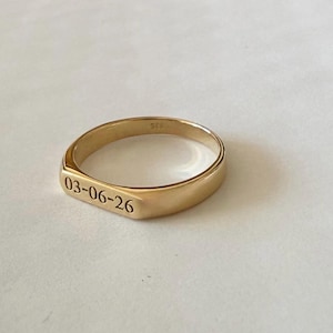 May include: A gold-toned band ring featuring a flat, rectangular front surface engraved with the date 03-06-26. The interior of the band displays a small 925 hallmark stamp, indicating the metal composition of this polished, smooth jewelry piece.