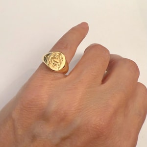 Peut inclure: Une bague chevalière en or avec un plateau ovale gravé d'un blason héraldique. La bague présente une finition polie et un anneau lisse et effilé. La gravure représente un écu avec trois éléments circulaires surmonté d'un casque et de volutes.
