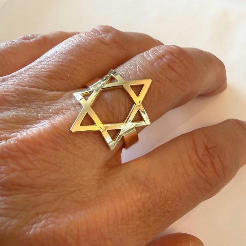 May include: A gold-colored ring featuring a Star of David design. The ring is worn on a finger and has a geometric shape. The Star of David is a six-pointed star, a symbol of Judaism. The ring is made of a shiny metal.