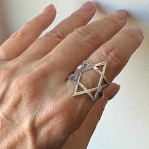 May include: A silver ring featuring a Star of David design. The ring is a geometric shape with six points, creating an open center. The ring is worn on a finger.