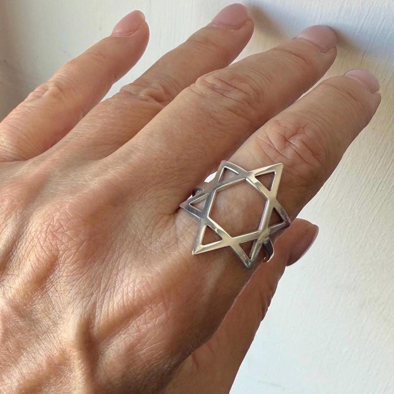 May include: A silver ring featuring a Star of David design. The ring is a geometric shape with six points, creating an open center. The ring is worn on a finger.